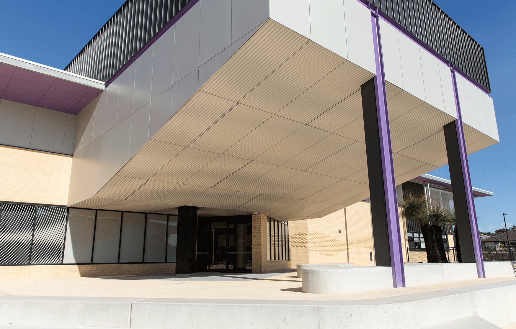 Looking up at the cantilevered college entry at Yanchep, its CNC routered soffit striped between purple columns.